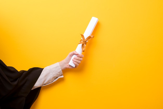 Close-up da mão da mulher em vestido de formatura está segurando o chapéu de formatura e certificado em fundo amarelo