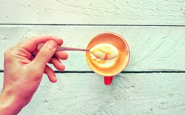 Close-up da mão de um barista levanta uma espuma com cappuccino fresco em um copo vermelho sobre uma mesa velha de madeira branca. Vista do topo