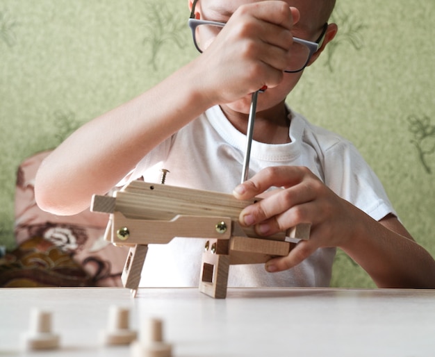 Close-up da mão de um menino monta um carrinho de brinquedo de madeira com uma chave de fenda sobre uma mesa de madeira em casa.