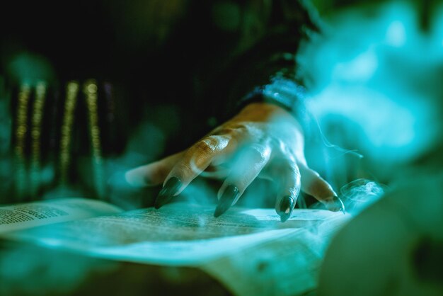 Close-up da mão de uma bruxa com unhas pretas pontiagudas no livro esfumaçado de receita mágica e com uma varinha mágica.