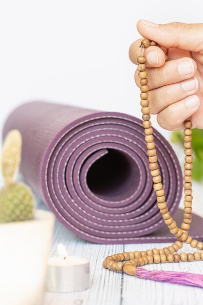 Close-up da mão de uma mulher, desdobrando um tapete de ioga violeta, contas de madeira ruins na madeira branca. acessórios essenciais para a prática de yoga e meditação.