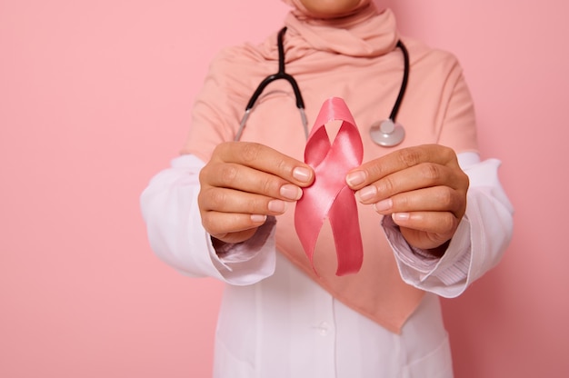 Close-up da mão do médico em hijab rosa, segurando a fita rosa. Dia Mundial do Câncer. Conscientização do câncer de mama e abdominal, dia rosa de outubro, fundo colorido, copie o espaço. Conceito de apoio ao câncer de mama.