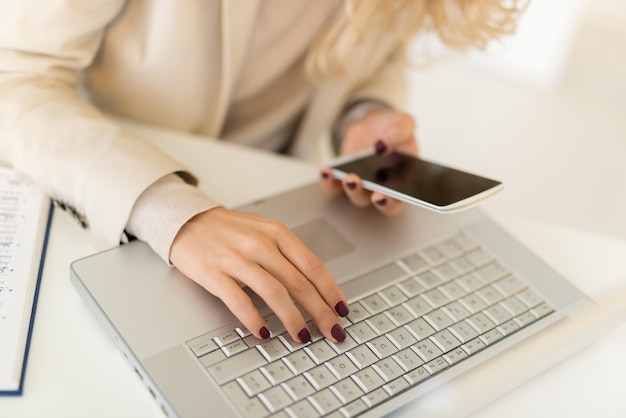 Close-up da mão feminina com unhas pintadas no teclado do laptop e digitando no telefone inteligente.