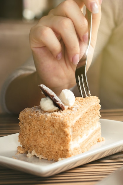 Foto close-up da mão feminina segurando o garfo de sobremesa sobre um pedaço de bolo de mel no café.