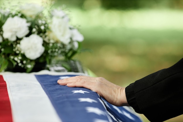 Foto close-up da mão masculina no caixão coberto com a bandeira americana, dizendo adeus na cerimônia fúnebre ao ar livre