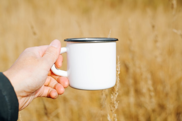 Close-up da mão mostrando a maquete da caneca branca em branco contra o campo de trigo amarelo ao ar livre. copo de viagem esmaltado com espaço vazio para logotipo ou marca. camping, caminhada, conceito de piquenique