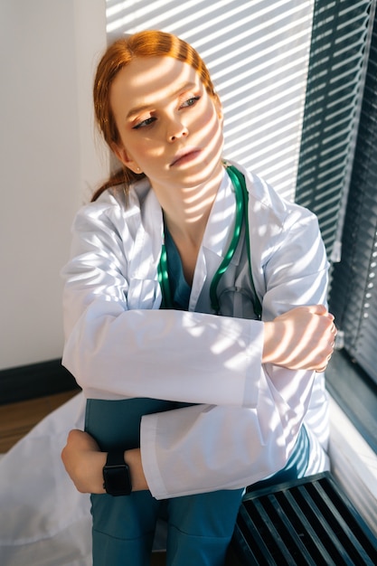 Close-up da médica jovem triste desesperada no jaleco branco, sentada no chão, abraçando as pernas com as mãos perto da janela.