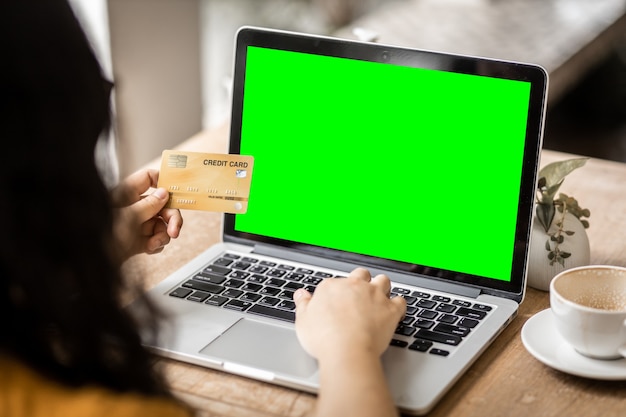 Close-up da mulher de negócios de pessoas autônomas Mão segurando os cartões de crédito casuais, trabalhando com um computador laptop com uma tela verde em branco na cafeteria como o plano de fundo, compras de pagamento on-line