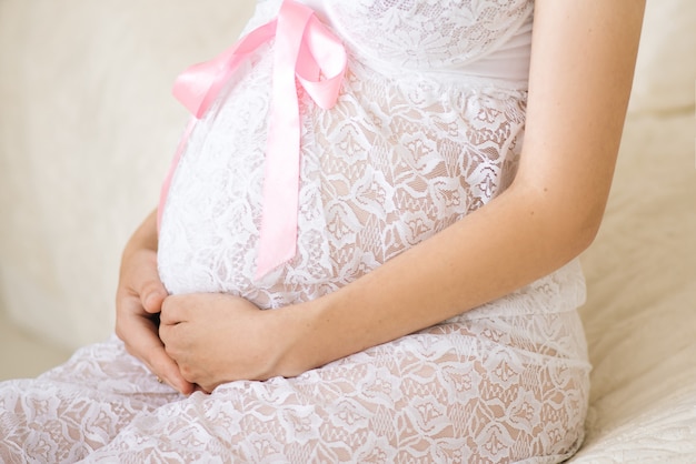 Close-up da mulher grávida irreconhecível com as mãos sobre a barriga em uma camisa de renda branca