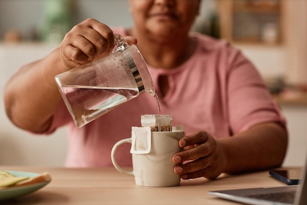 Close-up da mulher negra sênior fazendo chá na cozinha caseira segurando a chaleira de vidro e derramando água