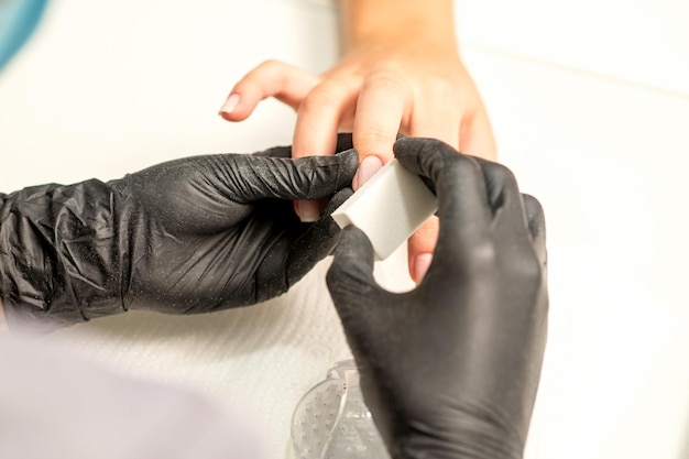 Close-up das mãos caucasianas de uma manicure profissional estão arquivando as unhas de uma jovem mulher caucasiana jovem recebendo uma manicure por uma esteticista com uma lixa de unha em um salão de beleza