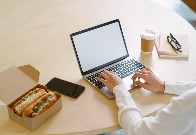 Close-up das mãos da mulher empreendedora trabalhando no computador portátil tem pausa para o almoço comendo sandwitch na mesa no escritório em casa no dia de inverno Quarentena de entrega de comida retirar comida Estilo de vida pouco saudável