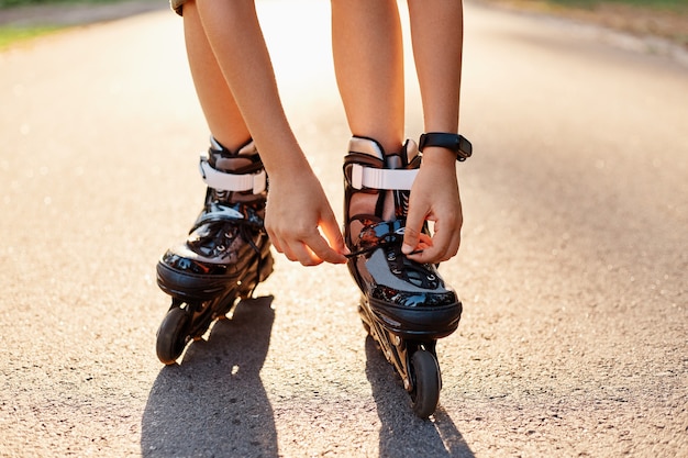 Close-up das mãos de uma criança anônima consertando cadarços de patins antes de patinar, criança desconhecida na estrada em um dia ensolarado de verão, andar de patins, estilo de vida ativo.