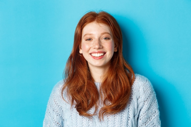 Close-up de alegre adolescente com cabelo ruivo cacheado, sorrindo feliz para a câmera, em pé contra um fundo azul.