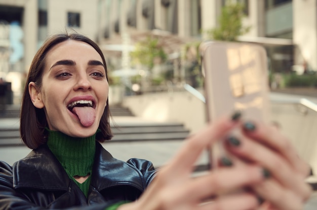 Foto close-up de alegre engraçada alegre mulher segurando o smartphone nas mãos estendidas e fazendo careta, mostrando a língua, tirando o autorretrato enquanto caminhava pela cidade. estilo de vida.