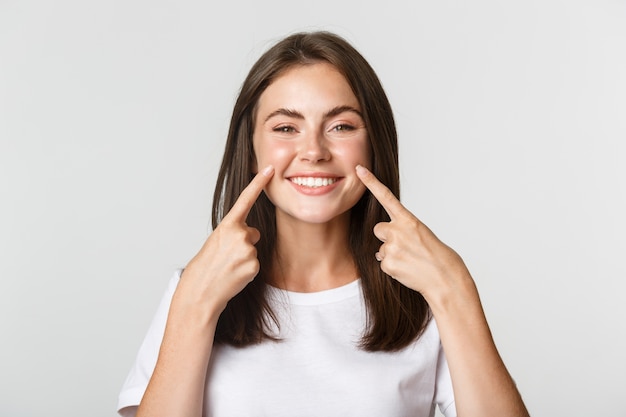 Foto close-up de atraente jovem sorridente apontando os dedos para o rosto, cutucando as bochechas, brancas.