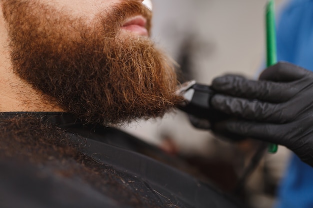 Close-up de cabeleireiro profissional atendendo cliente com barba espessa por tosquiadeira