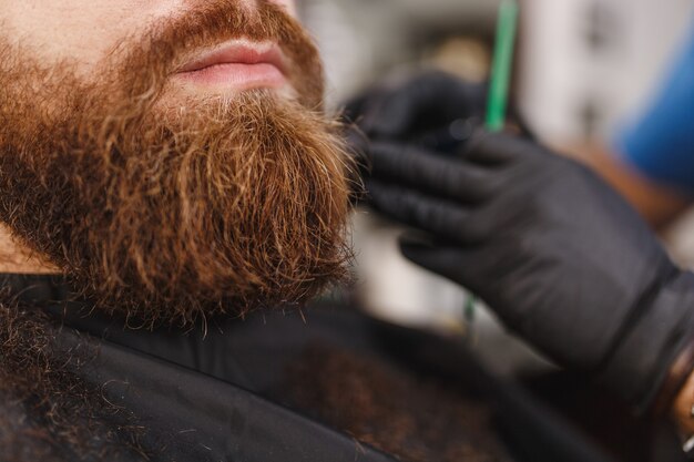 Close-up de cabeleireiro profissional atendendo cliente com barba espessa por tosquiadeira