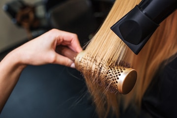 Cabeleireiro secando cabelos loiros à mão com secador de cabelo e escova  redonda em um salão de beleza.