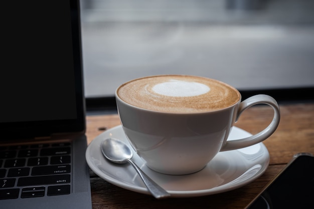 Close-up de café com leite quente com espuma de leite latte art na caneca do copo e computador laptop e smartphone na mesa de madeira da mesa de escritório no café no café, durante o conceito de trabalho de negócios