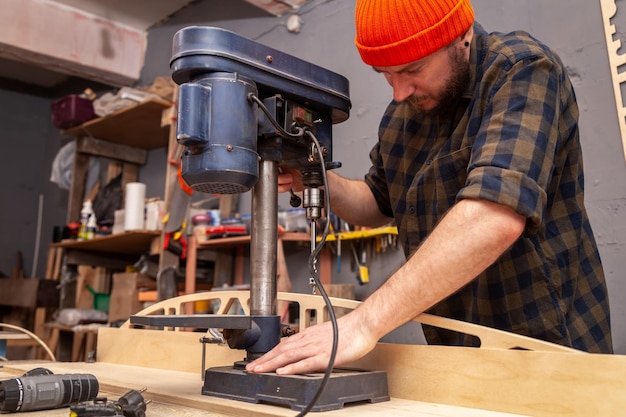 Close-up de carpinteiro experiente em roupas de trabalho e pequeno empresário está esculpindo uma placa de madeira em uma broca de mão moderna em uma visão lateral de oficina leve no fundo muitas ferramentas