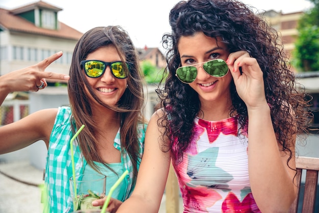 Close-up de duas mulheres jovens felizes com óculos de sol, sorrindo e olhando para a câmera, segurando smoothies de vegetais verdes ao ar livre