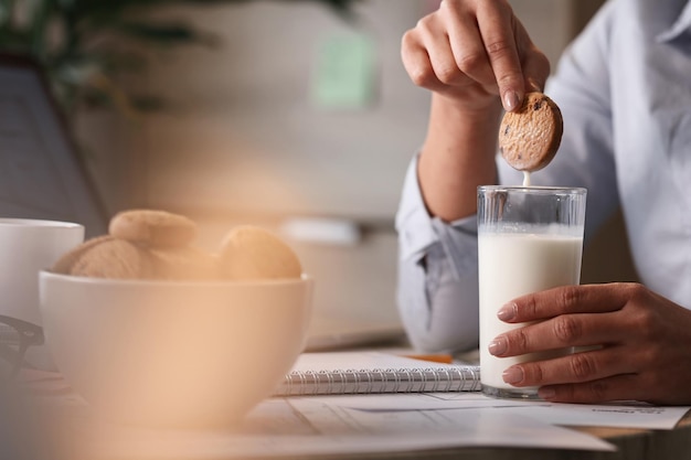 Close-up de empresária fazendo um lanche no trabalho e mergulhando biscoito em um copo de leite