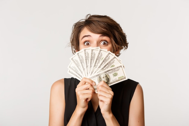 Close-up de excitada garota feliz segurando dinheiro perto do rosto e espiando para a câmera emocionado, em pé sobre o branco.