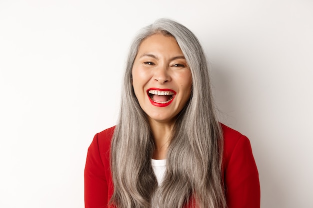 Close-up de feliz empresária asiática com longos cabelos grisalhos, vestindo um blazer vermelho, rindo e sorrindo alegremente para a câmera, fundo branco