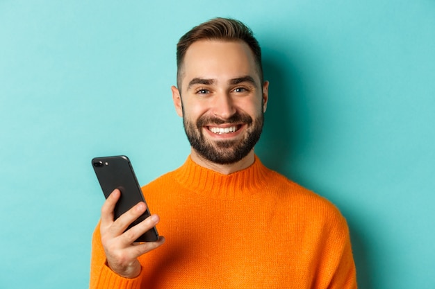 Close-up de feliz homem bonito escrevendo mensagem no celular, segurando o smartphone e sorrindo, de pé contra o fundo turquesa.