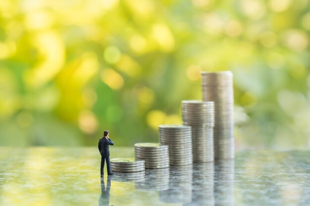 Close-up de figuras em miniatura de empresário em pé e olhando tp pilha de moedas com natureza de folha verde bokeh