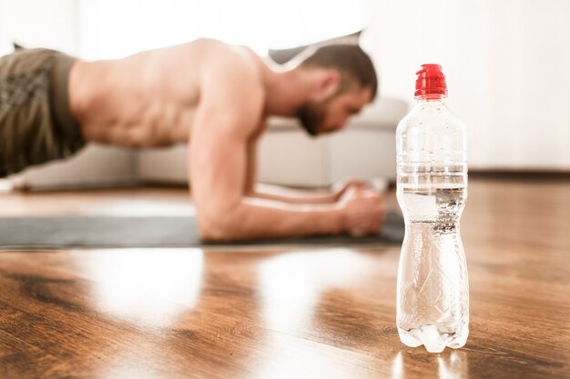 Close-up de garrafa de água. jovem fazendo esporte em casa. corte a vista do esportista de cara sem camisa com uma figura de esportes em posição de prancha.