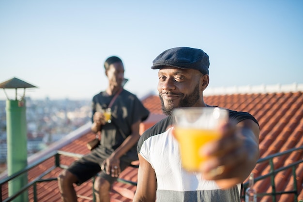 Close-up de homem Africano feliz segurando o copo de suco de fruta. Jovem barbudo de boné preto em pé no telhado sorrindo esticando a mão com um copo de suco para a câmera. Conceito de descanso e relações humanas