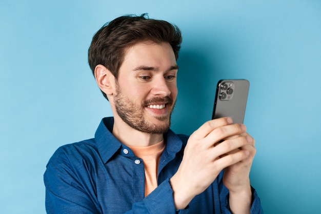 Close-up de homem bonito caucasiano escrevendo mensagem, lendo a tela do celular e sorrindo, de pé sobre um fundo azul.