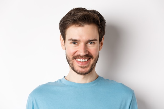 Close-up de homem bonito caucasiano sorrindo com dentes brancos, olhando confiante para a câmera, em pé de camisa azul sobre fundo branco.