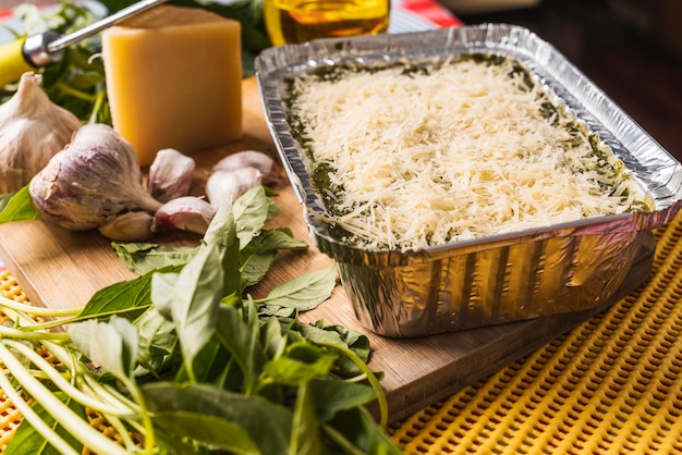 Close-up de lasanha caseira com pesto em uma bandeja de alumínio com bastante queijo ralado em cima em uma placa de madeira