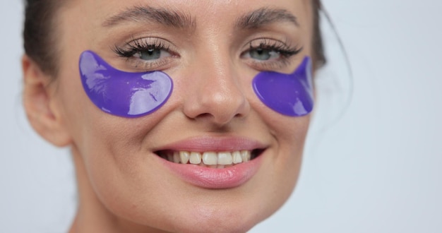 Foto close-up de linda mulher sorridente aplicando tapa-olhos violeta com confiança olhando na câmera sobre fundo cinza conceito de beleza