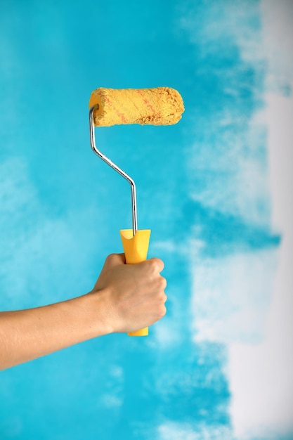 Foto close-up de mão feminina segurando rolo amarelo de tinta sobre construção de reparo de fundo azul