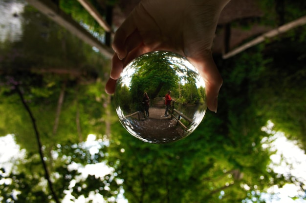 Close-up de mão segurando bola de cristal com reflexo de árvores ponte e 2 pessoas em Dulwich Park