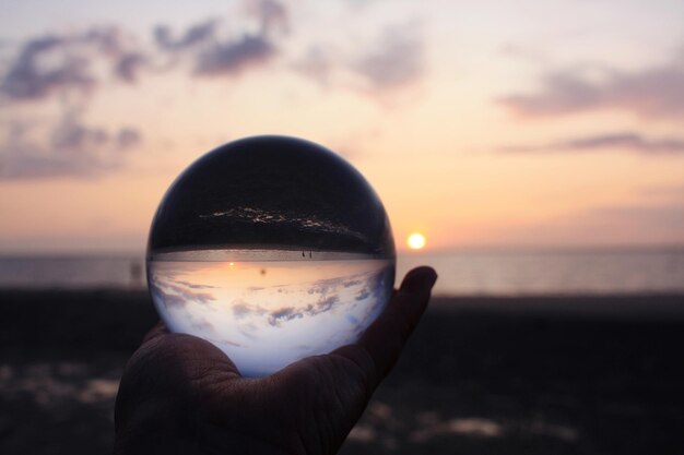 Close-up de mão segurando bola de cristal contra o mar durante o pôr do sol