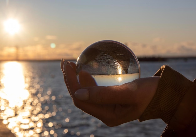 Foto close-up de mão segurando bola de cristal contra o mar durante o pôr do sol