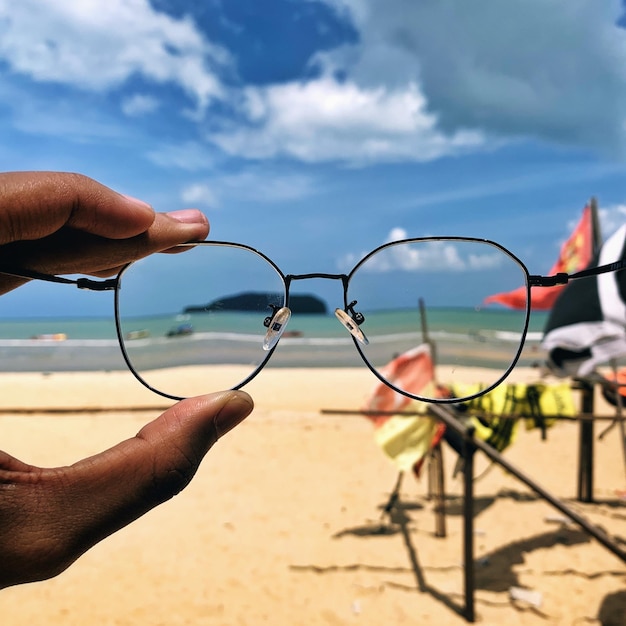 Foto close-up de mão segurando óculos na praia contra o céu