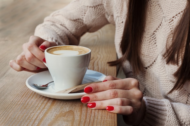 Close-up de mãos femininas com manicure de esmalte de gel vermelho segurando xícara com café