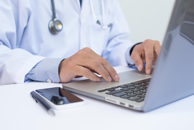 Close-up de médico masculino trabalhando em computador portátil e smartphone móvel em mesa branca em sistema de registros eletrônicos de saúde EHRs on-line médico ou conceito de telemedicina