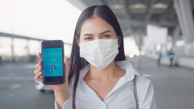 Close-up de mulher de negócios está usando máscara protetora no aeroporto internacional, mostrando passaporte de vacina em seu smartphone