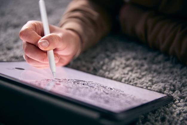 Close-up de mulher desenhando em tablet digital usando caneta stylus deitado no tapete em casa