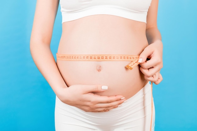 Close-up de mulher grávida em cueca branca medindo sua barriga em crescimento com uma fita métrica no fundo azul Verifique o desenvolvimento do bebê Copiar espaço