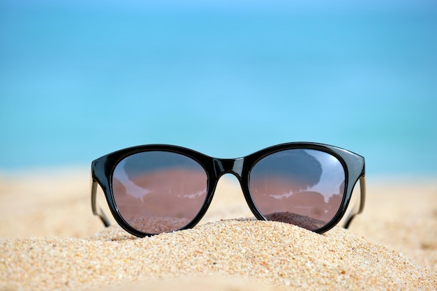 Close-up de óculos de sol protetores pretos na praia de areia à beira-mar tropical em dia de sol quente. Conceito de férias de verão.