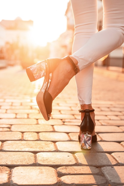 Close-up de pernas femininas em sandálias de couro pretas da moda de verão em jeans elegantes em uma estrada de pedra em um dia ensolarado