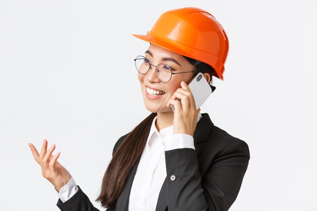 Close-up de profissional sorridente empresário asiático feminino na fábrica, engenheiro-chefe em capacete de segurança e terno, falando ao telefone, tendo uma conversa de negócios com investidores da empresa